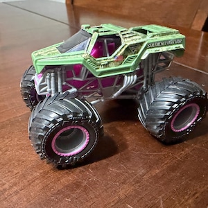 May include: A green and pink toy monster truck with large black tires and the words "SOLDIER FORTUNE" printed on the top. The truck has a camouflage design on the top and pink accents.