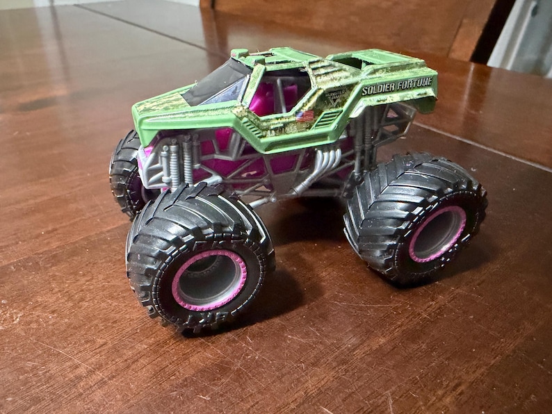 May include: A green and pink toy monster truck with large black tires and the words "SOLDIER FORTUNE" printed on the top. The truck has a camouflage design on the top and pink accents.