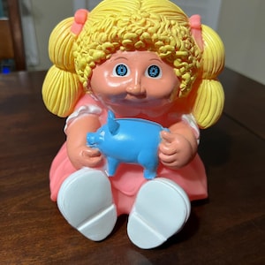 Puede incluir: Una figura de muñeca Cabbage Patch Kids vintage, con cabello rubio con lazos rosas, ojos azules y un vestido rosa. La muñeca sostiene una figura de cerdo azul. Los pies de la muñeca son blancos.
