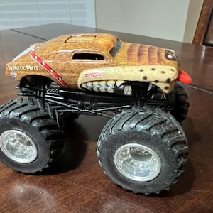 May include: A toy Monster Mutt monster truck with a brown and tan dog-like design. The truck has large black tyres, silver rims, and the Hot Wheels logo. The truck has a red tongue sticking out.