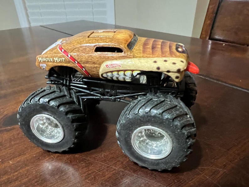 May include: A toy Monster Mutt monster truck with a brown and tan dog-like design. The truck has large black tyres, silver rims, and the Hot Wheels logo. The truck has a red tongue sticking out.