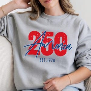 Puede incluir: Sudadera gris claro con el texto "250 America EST. 1776" en rojo y azul. La sudadera es de cuello redondo y tiene mangas largas.