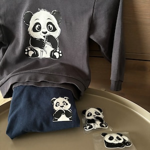 Puede incluir: Una sudadera gris oscuro con un gráfico de panda blanco y negro. El panda está sosteniendo un tallo de bambú en su boca. La sudadera está doblada y colocada encima de una camiseta azul oscuro con un gráfico de panda más pequeño. Hay dos pequeñas pegatinas de panda en una superficie blanca.