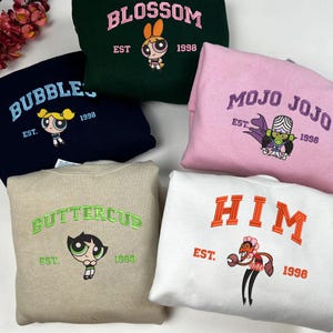 Könnte beinhalten: Eine Auswahl an Sweatshirts in verschiedenen Farben, jeweils mit einem gestickten Powerpuff Girls-Charakter und -Namen. Die Sweatshirts zeigen Blossom, Bubbles, Buttercup, Mojo Jojo und Ihn, mit dem Jahr 1998.