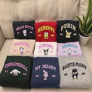 Puede incluir: Una colección de sudaderas coloridas con varios personajes de dibujos animados y texto. Cada sudadera muestra el nombre de un personaje, el año 1989 y la palabra «EST». Los colores incluyen negro, rojo, beige, blanco, morado, verde, azul marino y gris.