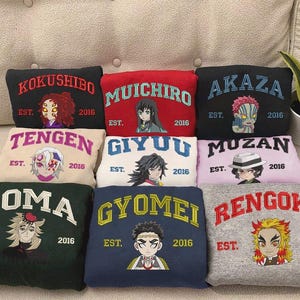 Könnte beinhalten: Eine Sammlung von bestickten Sweatshirts in verschiedenen Farben, jeweils mit einem Charakter-Namen und einer Illustration sowie dem Text "EST. 2016". Die Sweatshirts sind auf einer hellen Oberfläche angeordnet.