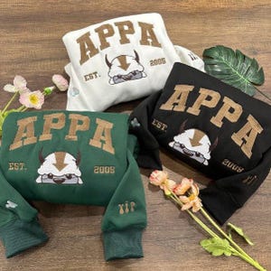 Könnte beinhalten: Drei Sweatshirts in Weiß, Schwarz und Grün, jeweils mit dem Wort "APPA" in braunen Blockbuchstaben bestickt. Darunter ein Cartoon-Charakter und das Jahr "2005" oder "2009". Die Sweatshirts sind auf einer Holzoberfläche gefaltet.