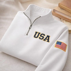 Könnte beinhalten: Ein weißes Sweatshirt mit einem Viertelreißverschluss-Kragen. Das Wort "USA" ist in Marineblau und Gold auf die Brust gestickt. Ein amerikanischer Flaggenaufnäher befindet sich auf dem Ärmel. Das Sweatshirt ist gefaltet.