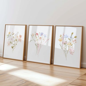 Puede incluir: Tres impresiones de acuarela enmarcadas de ramos de flores con flores rosas, azules y amarillas. Las impresiones están en marcos dorados simples y están colgadas en una pared blanca.