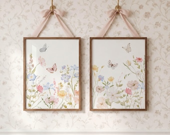 Puede incluir: Dos cuadros rectangulares con marco de madera que muestran ilustraciones en acuarela de flores silvestres en tonos rosa, azul y amarillo con mariposas. Cada marco está colgado de una cinta rosa claro atada con un lazo en un gancho.