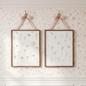 May include: Two rectangular wooden frames containing white prints with delicate watercolor illustrations of various wildflowers in soft pastel shades of pink, blue, and yellow. Each frame is suspended by a light pink ribbon tied in a bow.