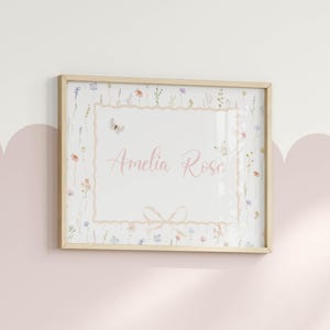 Può includere: Una stampa incorniciata con il nome "Amelia Rose" in una scrittura delicata. L'opera d'arte è circondata da un bordo floreale, farfalle e un fiocco. La cornice è in legno chiaro, aggiungendo un tocco di calore.