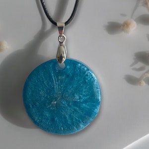 Peut inclure: Un pendentif rond en résine bleue avec des paillettes argentées, suspendu à un collier en cordon noir.