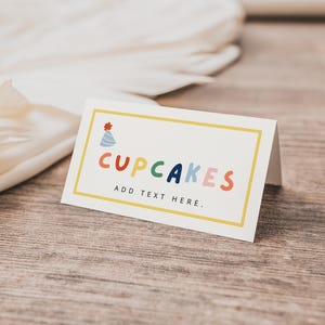 May include: A rectangular card with the word "CUPCAKES" in colorful letters, a party hat icon, and the text "ADD TEXT HERE." The card has a yellow border and is on a wooden surface.
