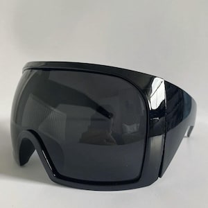 Puede incluir: Gafas de sol negras de estilo futurista con una montura ancha y curvada. Las lentes son oscuras y la montura tiene un acabado brillante. El diseño es elegante y moderno, adecuado para varios estilos de moda.