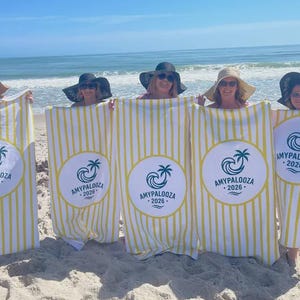 May include: Beach towels with yellow and white stripes and a circular logo that says "AMYPALOOZA - 2026". The towels are held up by people on a sandy beach with the ocean in the background.