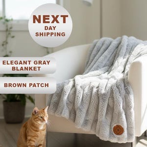 May include: An elegant gray blanket draped over a white armchair. The blanket has a textured, plush appearance. A brown patch is sewn onto the blanket. A ginger cat sits on the floor in front of the chair.