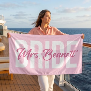 May include: A light pink beach towel with the word "BRIDE" in large white letters and "Mrs. Bennett" in a cursive pink font. The towel is held up on a cruise ship deck with the ocean in the background.
