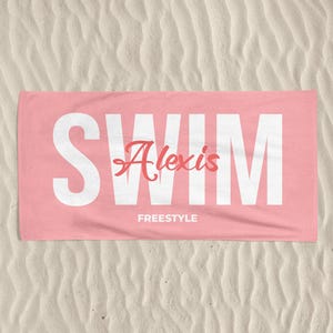 May include: A pink beach towel with the word "SWIM" in large white capital letters. The name "Alexis" is written in a cursive font in the center, with "FREESTYLE" below it. The towel is lying on a sandy surface.