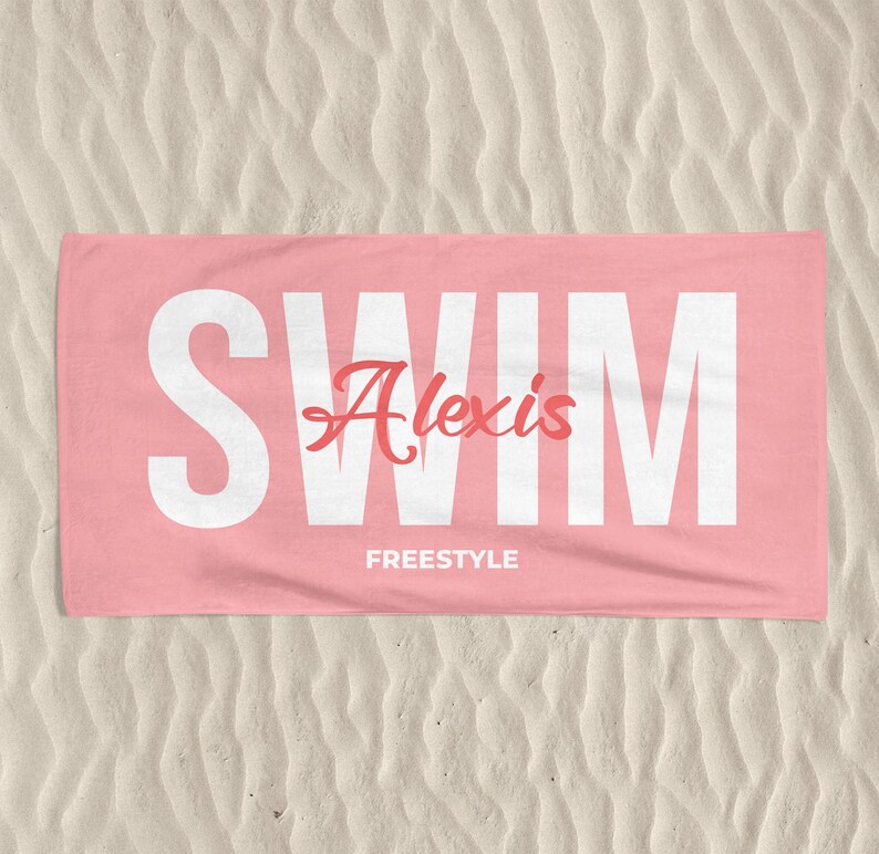 May include: A pink beach towel with the word "SWIM" in large white capital letters. The name "Alexis" is written in a cursive font in the center, with "FREESTYLE" below it. The towel is lying on a sandy surface.