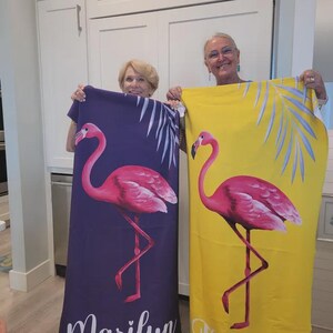 May include: Two beach towels, one purple and one yellow, each featuring a pink flamingo design. The purple towel has the name "Marilyn" printed at the bottom. Both towels have a tropical theme with palm fronds.