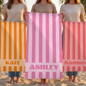 May include: Three beach towels with vertical stripes in orange, pink, and coral colors. Each towel has a name printed at the bottom: "KATE", "ASHLEY", and "MADISON". The towels are held up by people on a sandy beach with palm trees.