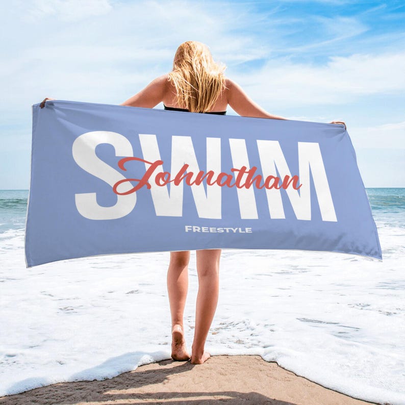 May include: A light blue beach towel with the word "SWIM" in large white letters. The name "Johnathan" is in a cursive red font, with the word "FREESTYLE" below. The towel is held up by a person on a beach.