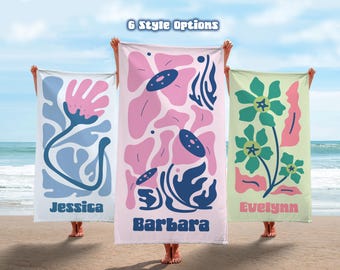 May include: Three beach towels with floral designs in pink, blue, and green are displayed on a beach. Each towel has a different floral pattern and a name printed on it. The text "6 Style Options" is at the top.