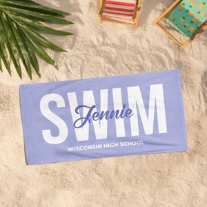 May include: A light purple beach towel with the word "SWIM" in large white letters. The name "Jennie" is written in cursive, and below it, "WISCONSIN HIGH SCHOOL" is printed in white. The towel is lying on sand, with two small beach chairs in the background.