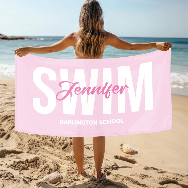 May include: A light pink beach towel with the word "SWIM" in large white letters. The name "Jennifer" is in pink script, and "DARLINGTON SCHOOL" is below. The towel is held up on a sandy beach with the ocean in the background.