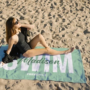 May include: A black swimsuit and sunglasses on a beach. A light green beach towel with the name "Madison" and the word "Swim" in large white letters is on the sand. The towel also has the word "Freestyle" printed on it.