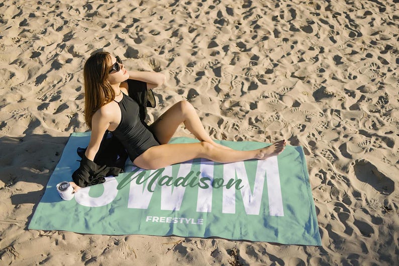 May include: A black swimsuit and sunglasses on a beach. A light green beach towel with the name "Madison" and the word "Swim" in large white letters is on the sand. The towel also has the word "Freestyle" printed on it.