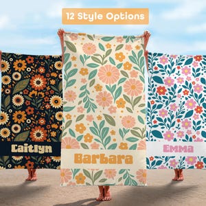 May include: Three beach towels with floral patterns are displayed on a beach. The towels are held up by people. The towels have different floral designs in various colors, including orange, pink, and blue. The text "12 Style Options" is at the top.