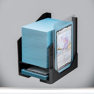Puede incluir: Soporte negro para cartas con una pila de cartas azules y una carta con texto. El soporte está montado en una pared gris. La tarjeta en el soporte tiene una imagen de una criatura y texto.
