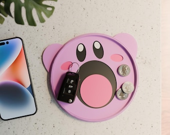 May include: A pink silicone tray with a cartoon face, featuring a large open mouth, two eyes, and two small cheeks. A black car key and three silver coins are placed inside the tray.