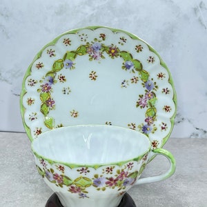 Pode incluir: Um conjunto de chávena e pires de porcelana branca com um design verde e floral. A chávena tem uma borda ondulada e o pires tem uma borda ondulada. O conjunto é decorado com pequenas flores rosa, roxas e amarelas.