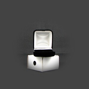May include: A black velvet ring box with a white interior is open and sitting on top of a white cardboard box.