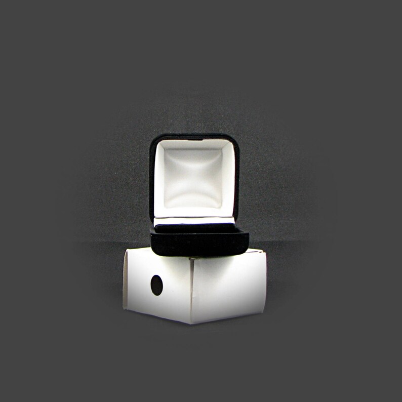 May include: A black velvet ring box with a white interior is open and sitting on top of a white cardboard box.