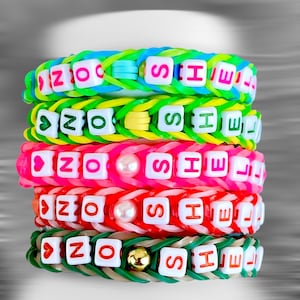 Può includere: Pila di braccialetti colorati in gomma. Ogni braccialetto è di un colore diverso, tra cui blu, verde, rosa, rosso e verde. I braccialetti hanno perline di lettere bianche che compongono il nome "NO SHELL".