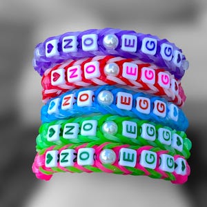 May include: A stack of colorful rubber band bracelets. Each bracelet is a different color, including purple, red, blue, green, and pink. The bracelets spell out the word "NO EGG" with white letter beads. Each bracelet has a pearl bead.