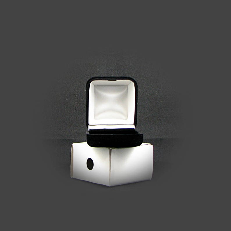 May include: A black velvet ring box with a white interior is open and sitting on top of a white cardboard box.