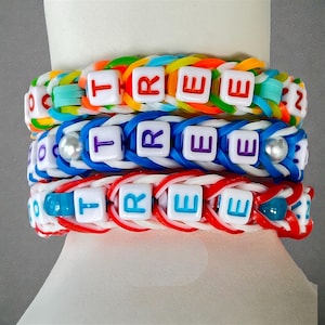 May include: Three colourful rubber band bracelets spelling the word "TREE". The bracelets are in rainbow, blue, and red colour schemes. Each bracelet has white letter beads spelling the word "TREE".