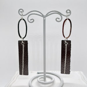 May include: A pair of brown leather earrings with silver oval hoops and a silver wire detail. The earrings are hanging from a white display stand.