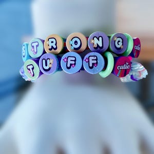 Op de afbeelding: Twee kleurrijke armbanden met de woorden "STRONG" en "TUFF" gespeld in ronde, pastelkleurige kralen. De armbanden hebben een mix van blauwe, paarse, groene en oranje tinten. Het woord "cutie" is ook zichtbaar op de armband.