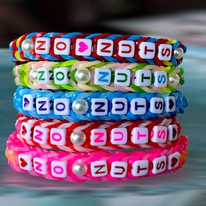 Op de afbeelding: Een stapel kleurrijke elastiekarmbanden. Elke armband heeft een andere kleur, waaronder rood, blauw, groen en roze. De armbanden spellen "NO NUTS" met witte letterkralen en hebben parelaccenten.