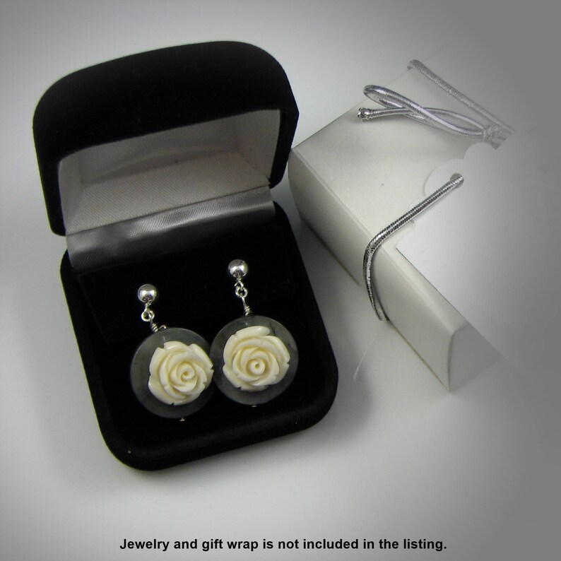 May include: A pair of white rose earrings in a black velvet jewelry box. The earrings are made of carved bone and have a silver tone finish.