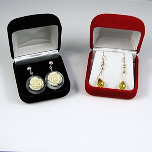 May include: Two velvet jewelry boxes, one red and one black, each containing a pair of earrings. The black box contains earrings with white flower-shaped pendants. The red box contains earrings with yellow teardrop-shaped pendants.