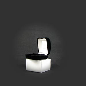 May include: A black velvet ring box with a white interior is open and sitting on a white box.
