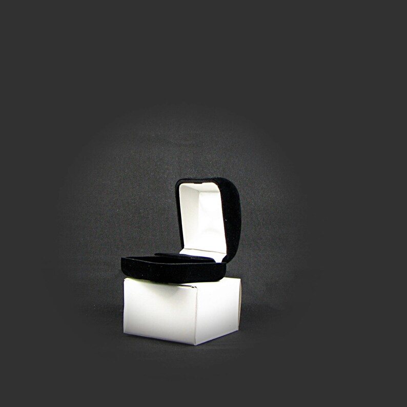 May include: A black velvet ring box with a white interior is open and sitting on a white box.