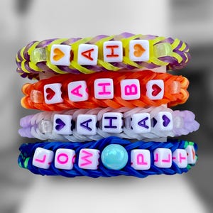 May include: Four colorful rubber band bracelets stacked on top of each other. Each bracelet is a different color: purple and yellow, orange, lavender, and blue. The bracelets have white lettered beads spelling out names and words.
